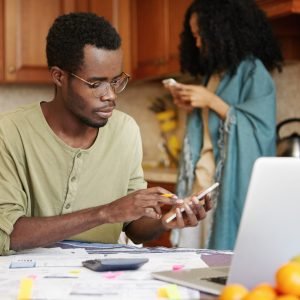 Dark-skinned man in spectacles doing finances, using cell phone, calculator and laptop computer, making call to utility services. Female with Afro hairstyle standing in background with mobile